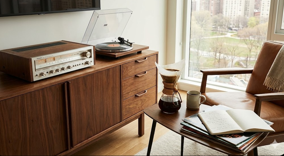 A sunlit modern living room with a vintage silver stereo receiver and turntable playing a record on a walnut cabinet. A Chemex coffee maker, mug, and stack of vinyl records are on a side table next to a leather armchair by a large window overlooking a park.