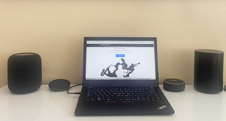 Four smart speakers sit on a white desk surrounding a black laptop displaying the Web Radio Info website. From left to right the speakers are an Apple HomePod, a Google Nest Mini, an Amazon Echo Dot, and a Sonos Era 100.