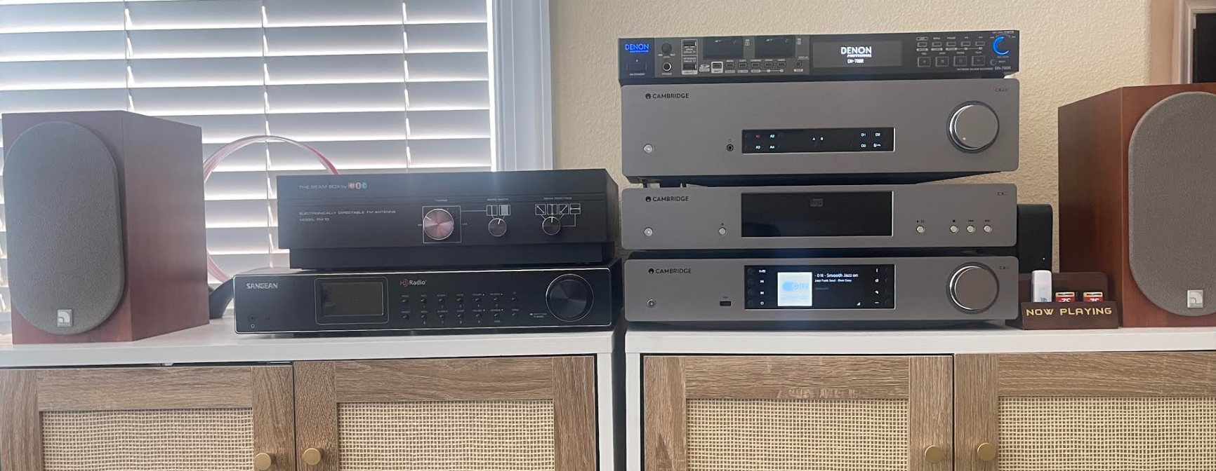 A detailed, photorealistic close-up photograph of a hand inserting a white 3.5mm auxiliary audio cable jack into the output port on the rear of a gray, spherical Amazon Echo Dot smart speaker. The speaker rests on a light wooden surface against a softly blurred, warm interior background. A small, circular blue and white "web radio info" logo is subtly visible on the gray fabric near the base. The depth of field is shallow, emphasizing the physical connection being made.