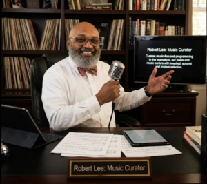 A smiling, bald Black man with a gray beard and glasses, wearing a white shirt and patterned bow tie, sits at a desk holding a vintage-style silver microphone. In front of him are scattered sheet music, two tablets, and a brass nameplate reading "Robert Lee: Music Curator." Behind him is a dark wood bookcase filled with vinyl records, books, and framed pictures, alongside a monitor displaying his name and title.